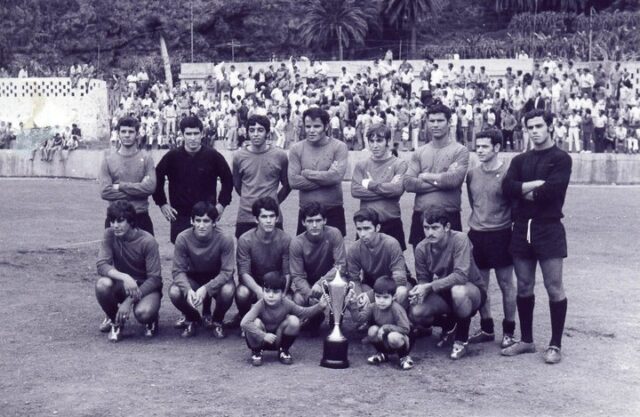 Equipo histórico del CD Mensajero junto a un trofeo, con la afición en las gradas